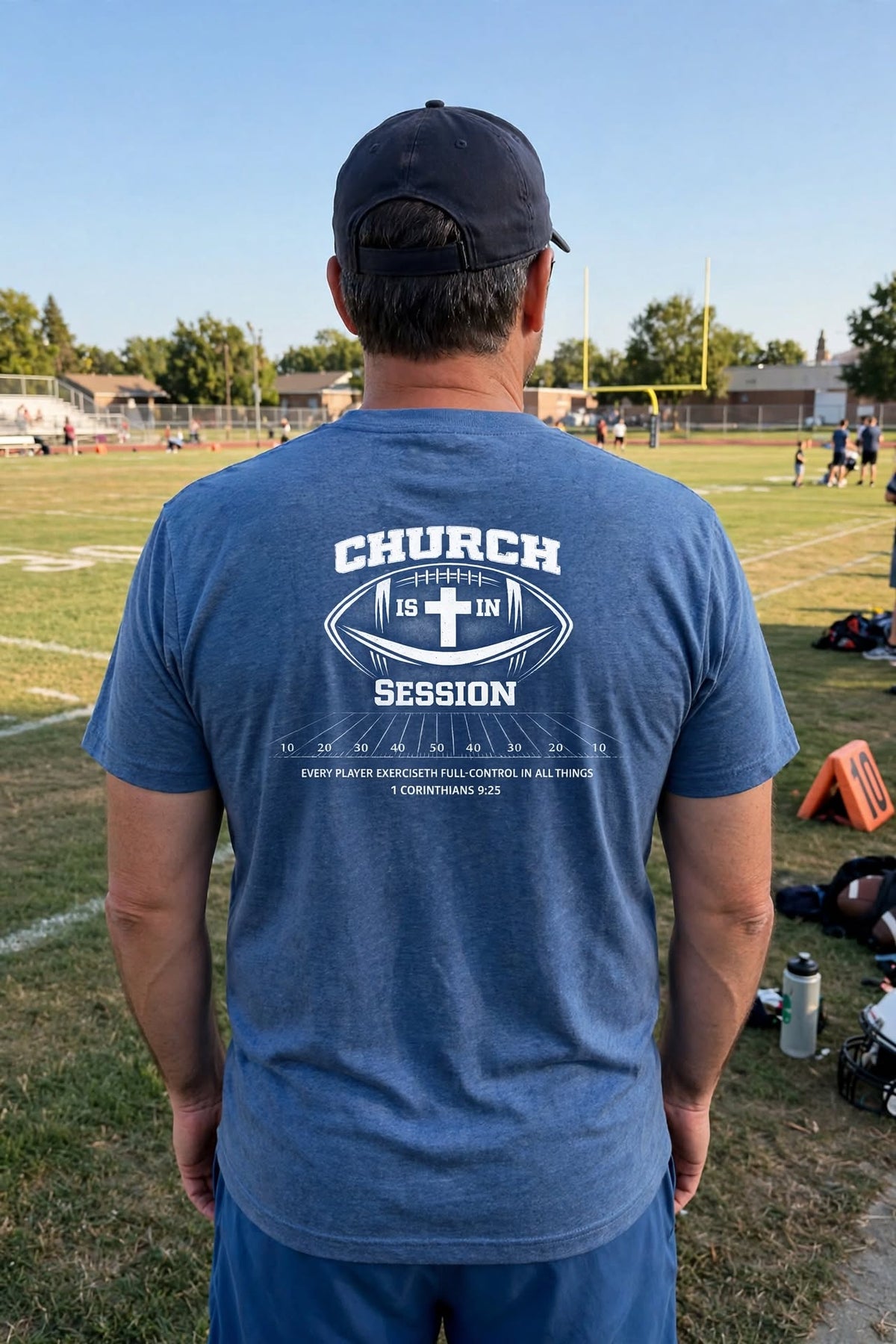 A man viewed from behind wearing a heathered blue t-shirt with a white graphic on the back that reads 'CHURCH SESSION' and a football motif, standing on a grassy sports field.