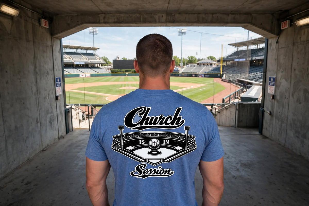 A man viewed from behind wearing a blue t-shirt with a large baseball graphic on the back that reads 'Church IS IN Season'. He is standing in a concrete tunnel looking out onto a baseball field.
